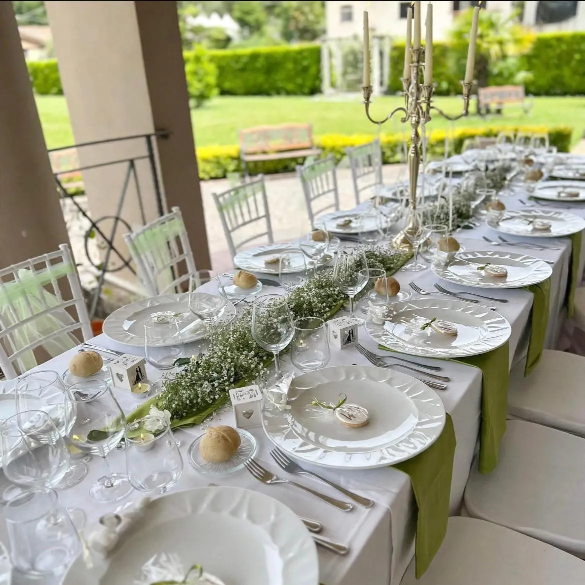 Tavola imperiale apparecchiata sotto il portico della villa, con candelabro d'argento, gypsophila e il giardino sullo sfondo